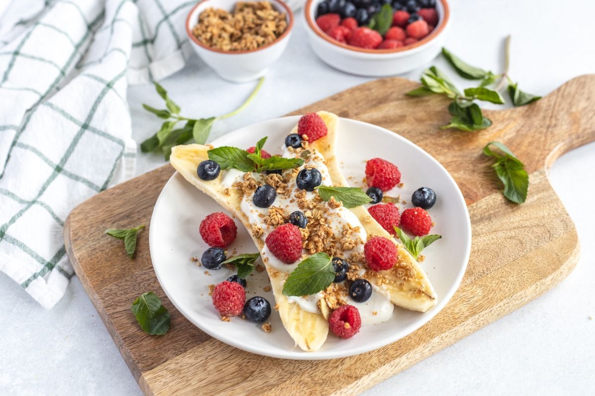 Breakfast Banana Split: A healthy breakfast idea for kids featuring yogurt, granola, raspberries, blueberries, and mint on a banana—all served on a white plate atop a wooden board. An easy no cook breakfast everyone will love!.