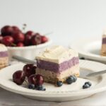 A plate with a layered No Bake Berry Layered Bars cheesecake made with raw cashews, topped with blueberries and cherries, and a fork beside it, with more cake and fruit in the background.