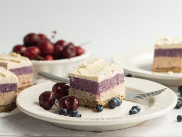 A plate with a layered No Bake Berry Layered Bars cheesecake made with raw cashews, topped with blueberries and cherries, and a fork beside it, with more cake and fruit in the background.
