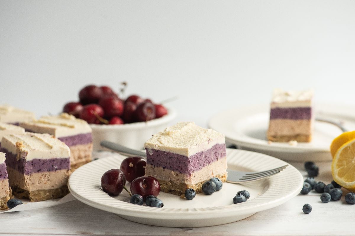 A slice of no bake vegan cheesecake made with raw cashews on a plate with blueberries and cherries, next to other dessert slices.