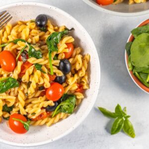 A bowl of easy spinach pasta with cherry tomatoes, black olives, and a fork beside a small bowl of fresh spinach.