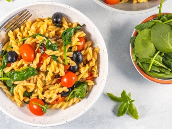 A bowl of easy spinach pasta with cherry tomatoes, black olives, and a fork beside a small bowl of fresh spinach.