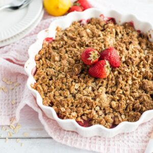 A strawberry crumble or crisp dessert in a white dish, topped with fresh strawberries, rests on a light pink cloth.