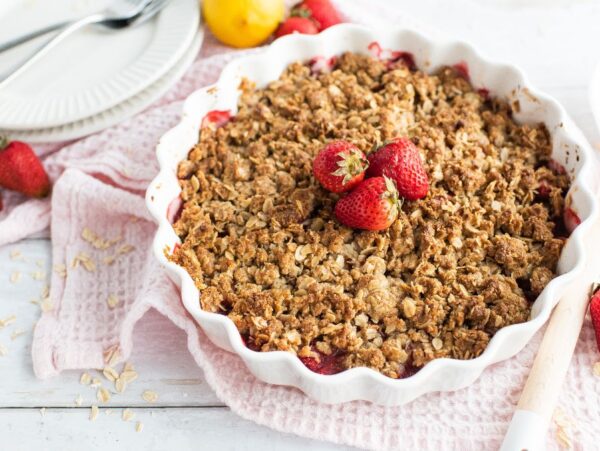 A strawberry crumble or crisp dessert in a white dish, topped with fresh strawberries, rests on a light pink cloth.