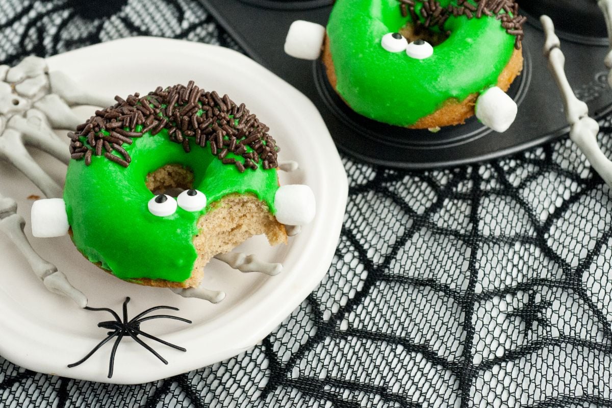 Green Frankenstein Apple Cider Halloween Donuts with candy eyes and chocolate sprinkles, on a skeleton hand plate with a spider. These baked apple cider donuts look like a Frankenstein monster—perfectly spooky for your Halloween celebration!.