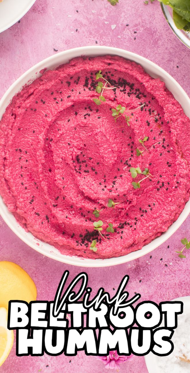 A bowl of vibrant pink beetroot hummus, topped with microgreens and black seeds, sits on a pink surface.