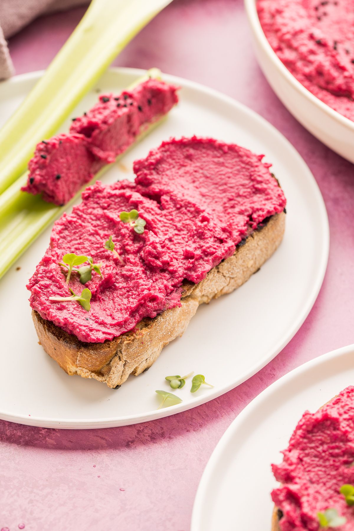 A slice of bread topped with vibrant pink beetroot hummus and garnished with microgreens on a white plate.