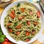 A plate of spaghetti with vegan pesto sauce, cherry tomatoes, and herbs sits on a yellow-checked tablecloth.