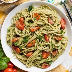 A plate of spaghetti with vegan pesto sauce, cherry tomatoes, and herbs sits on a yellow-checked tablecloth.