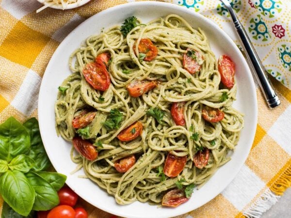 A plate of spaghetti with vegan pesto sauce, cherry tomatoes, and herbs sits on a yellow-checked tablecloth.