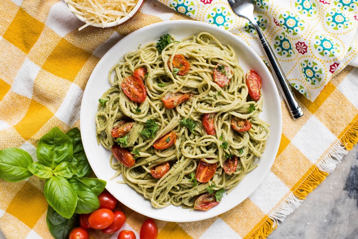 A bowl of spaghetti with vegan pesto sauce, cherry tomatoes, and parsley on a checkered tablecloth, with basil and cheese nearby.