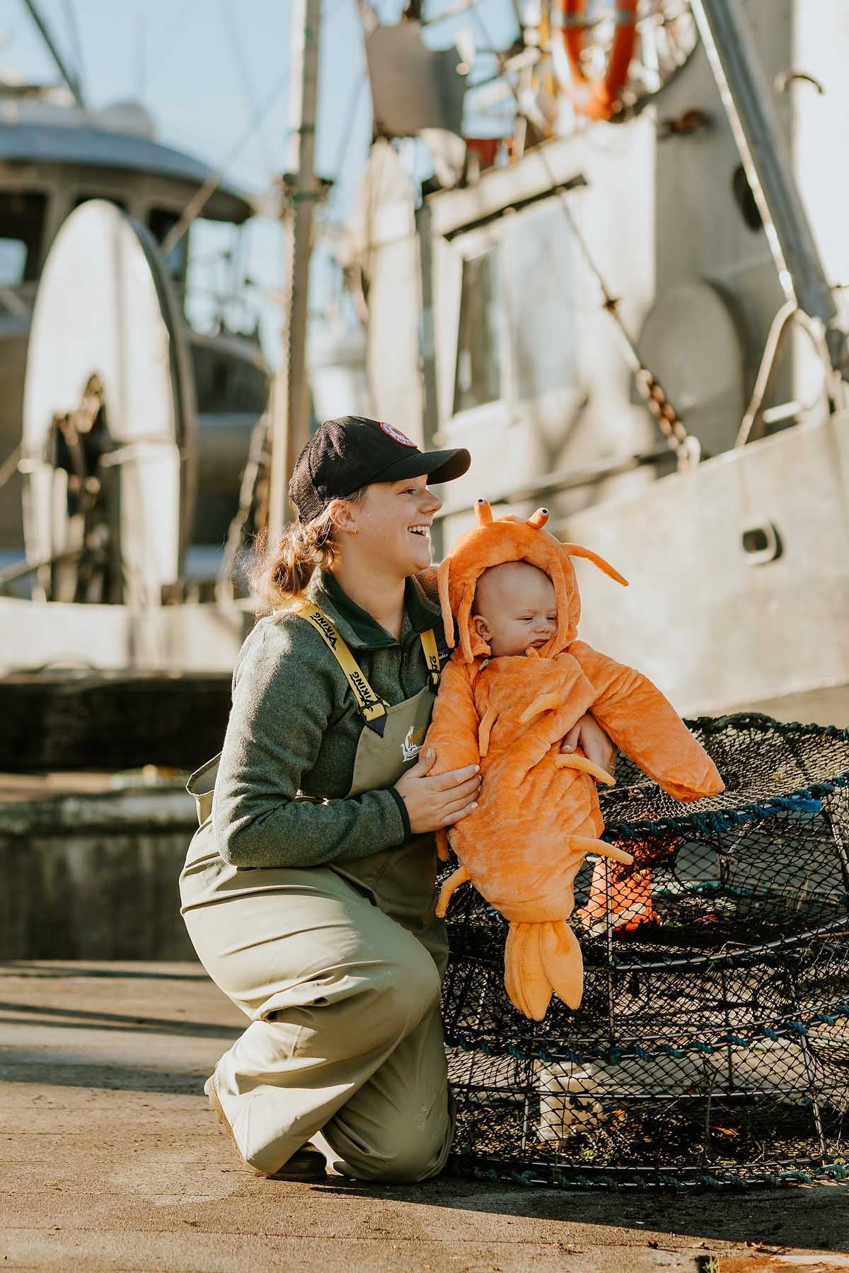 A person kneels beside a baby in an orange lobster costume near fishing boats and nets at a dock.