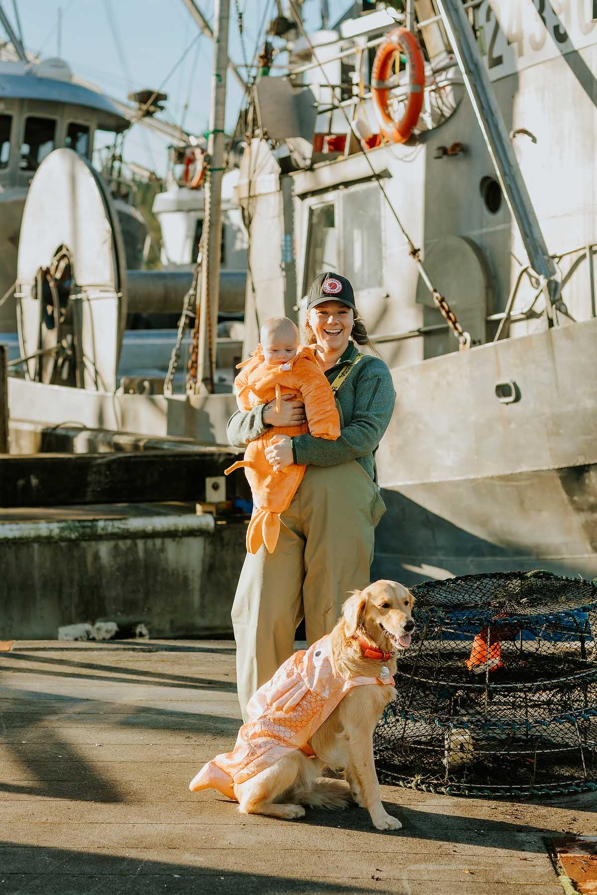 A person holds a baby, both in orange outfits, with a dog in costume by fishing boats and nets on a dock.