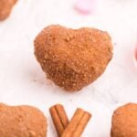 Heart-shaped cinnamon sugar pastries made with a red heart cookie cutter and cinnamon sticks—try this mini doughnut recipe for a sweet cinnamon roll hack on a white surface.
