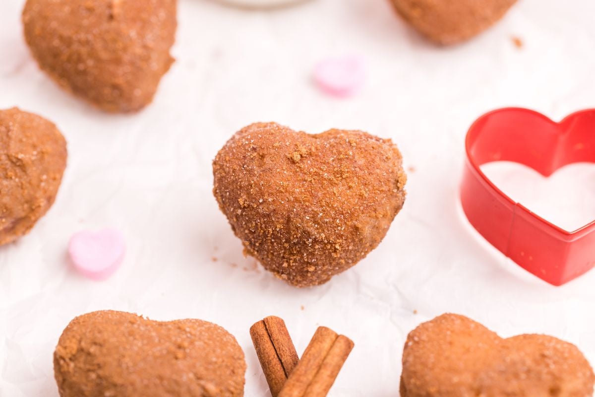 Heart-shaped donut covered in cinnamon sugar, surrounded by candy hearts and a red heart-shaped cookie cutter—perfect inspiration for your next mini doughnut recipe.