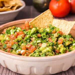 A bowl of chunky guacamole made from an easy guacamole recipe sits with tortilla chips, surrounded by fresh tomatoes, avocado, and cilantro.