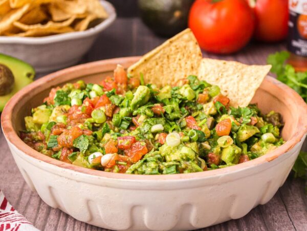 A bowl of chunky guacamole made from an easy guacamole recipe sits with tortilla chips, surrounded by fresh tomatoes, avocado, and cilantro.