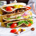 Three stacked tacos filled with ground meat, lettuce, tomato, corn, onion, and cheese showcase the popular folded tortilla hack on a white surface.