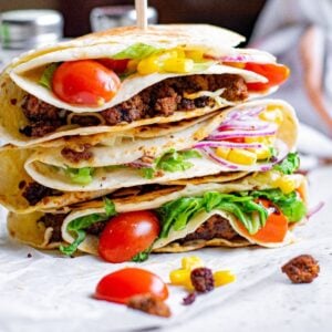 Three stacked tacos filled with ground meat, lettuce, tomato, corn, onion, and cheese showcase the popular folded tortilla hack on a white surface.