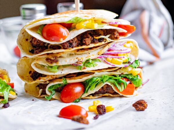 Three stacked tacos filled with ground meat, lettuce, tomato, corn, onion, and cheese showcase the popular folded tortilla hack on a white surface.