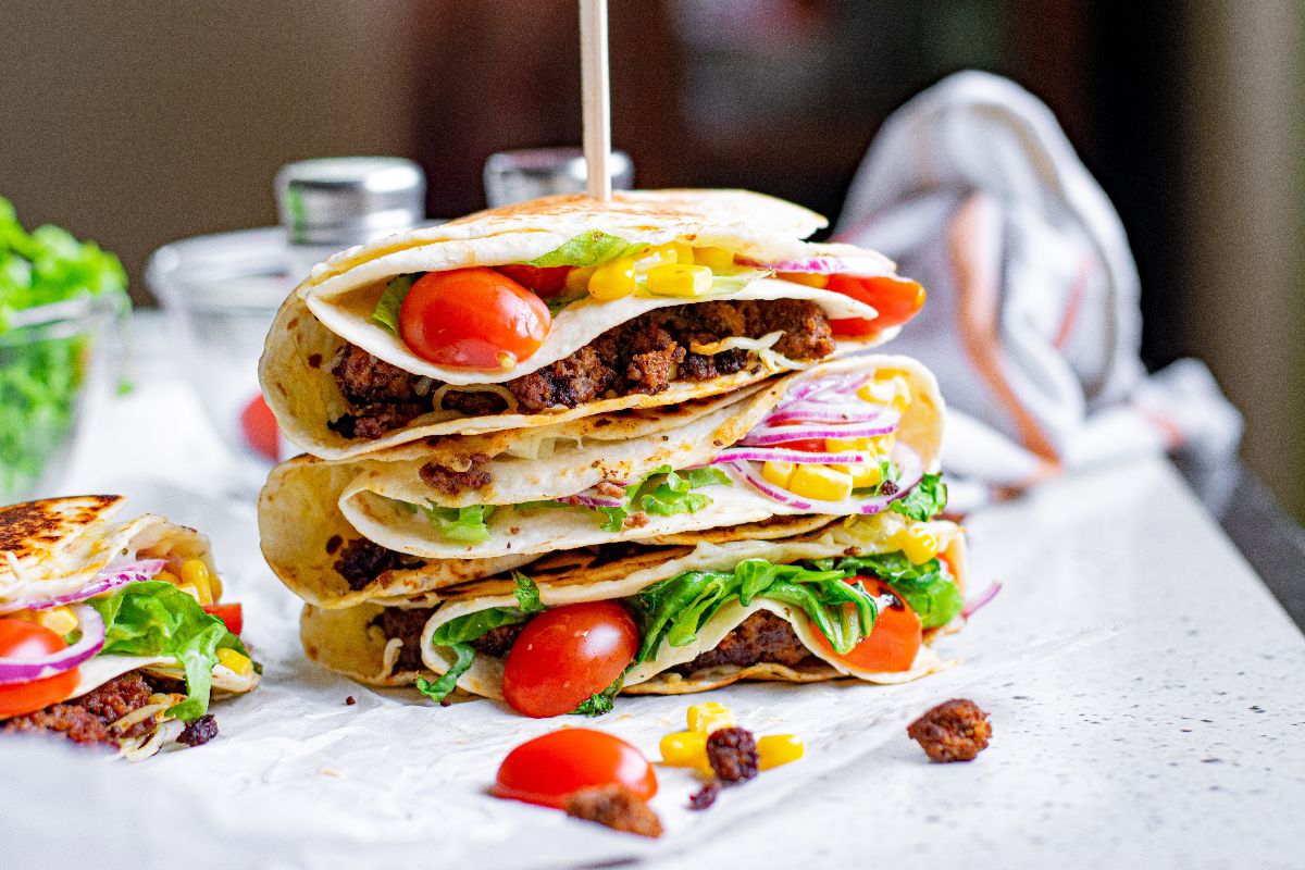 Three stacked tacos filled with ground meat, lettuce, tomatoes, onions, and corn on a countertop—reminiscent of the viral TikTok folded tortilla trend.