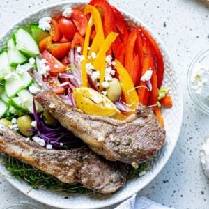 A bowl with grilled lamb chops, sliced cucumber, tomato, bell pepper, olives, and feta cheese on a white surface—perfect for pairing with a Greek lemon baked pork chops recipe.