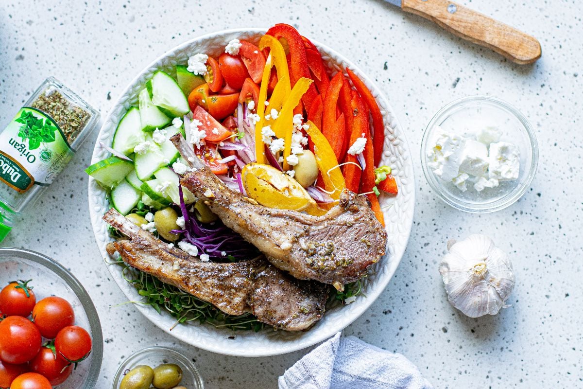 A bowl with grilled lamb chops or Greek pork chops, sliced veggies, olives, feta cheese, and herbs on a light countertop.