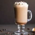 Glass mug of creamy Irish cream coffee topped with whipped cream and chocolate shavings, with a spoon and coffee beans nearby.