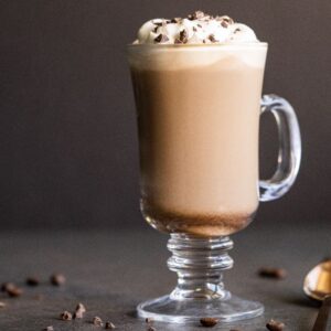 Glass mug of creamy Irish cream coffee topped with whipped cream and chocolate shavings, with a spoon and coffee beans nearby.