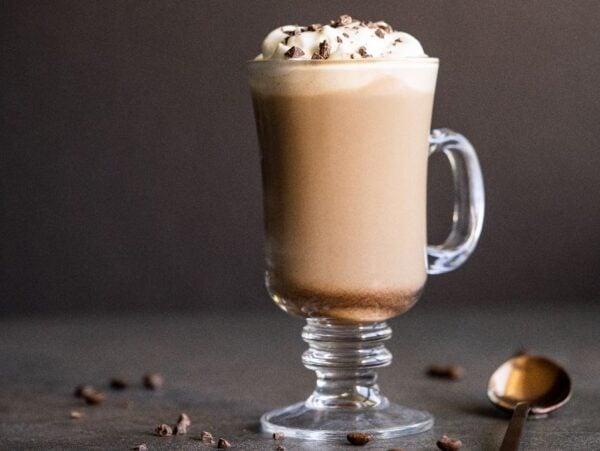 Glass mug of creamy Irish cream coffee topped with whipped cream and chocolate shavings, with a spoon and coffee beans nearby.