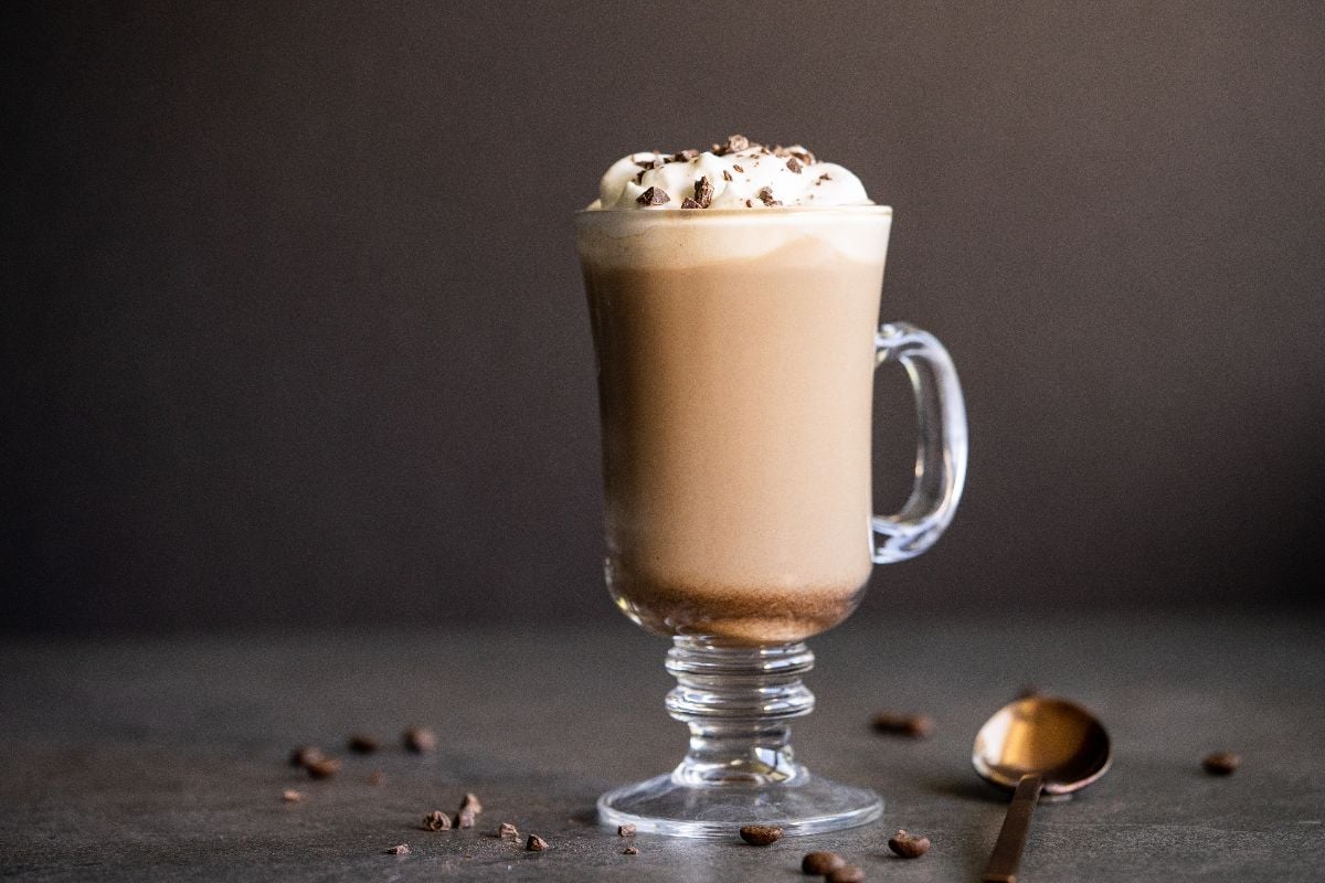A glass mug of Irish cream coffee topped with whipped cream and chocolate shavings, with coffee beans and a spoon nearby.