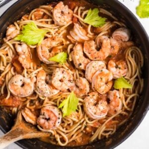 Spicy shrimp pasta with Bloody Mary vodka sauce, tomato, and celery leaves in a black pot, served with a wooden spoon on the side.