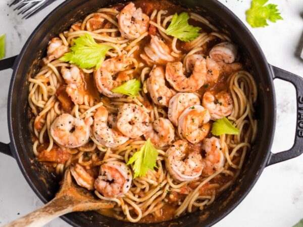 Spicy shrimp pasta with Bloody Mary vodka sauce, tomato, and celery leaves in a black pot, served with a wooden spoon on the side.