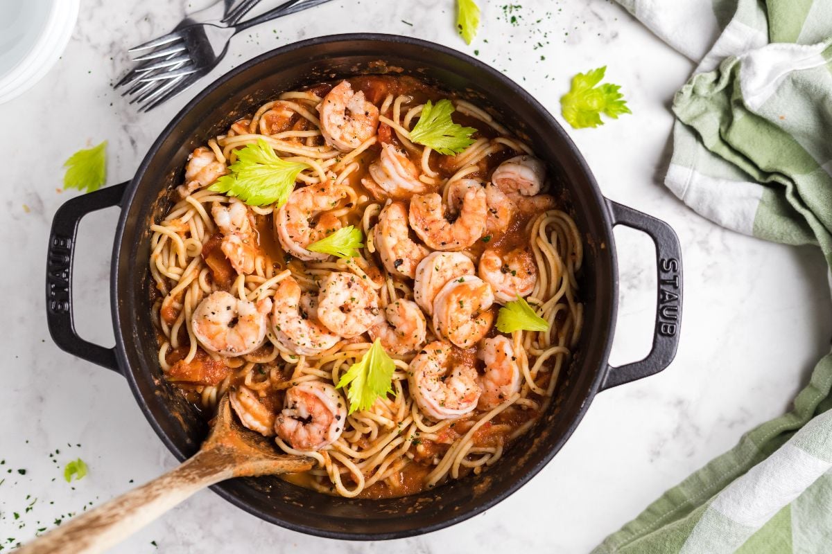 A pot of spicy shrimp pasta with tomato sauce, garnished with celery leaves, sits on a marble surface.
