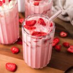 Three glasses of pink strawberry milkshake with glass straws, topped with fresh strawberry slices on a wooden board—perfect for a homemade Starbucks pink drink vibe.