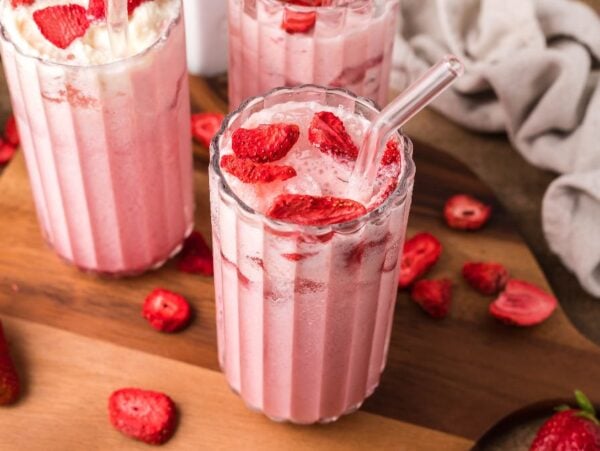Three glasses of pink strawberry milkshake with glass straws, topped with fresh strawberry slices on a wooden board—perfect for a homemade Starbucks pink drink vibe.