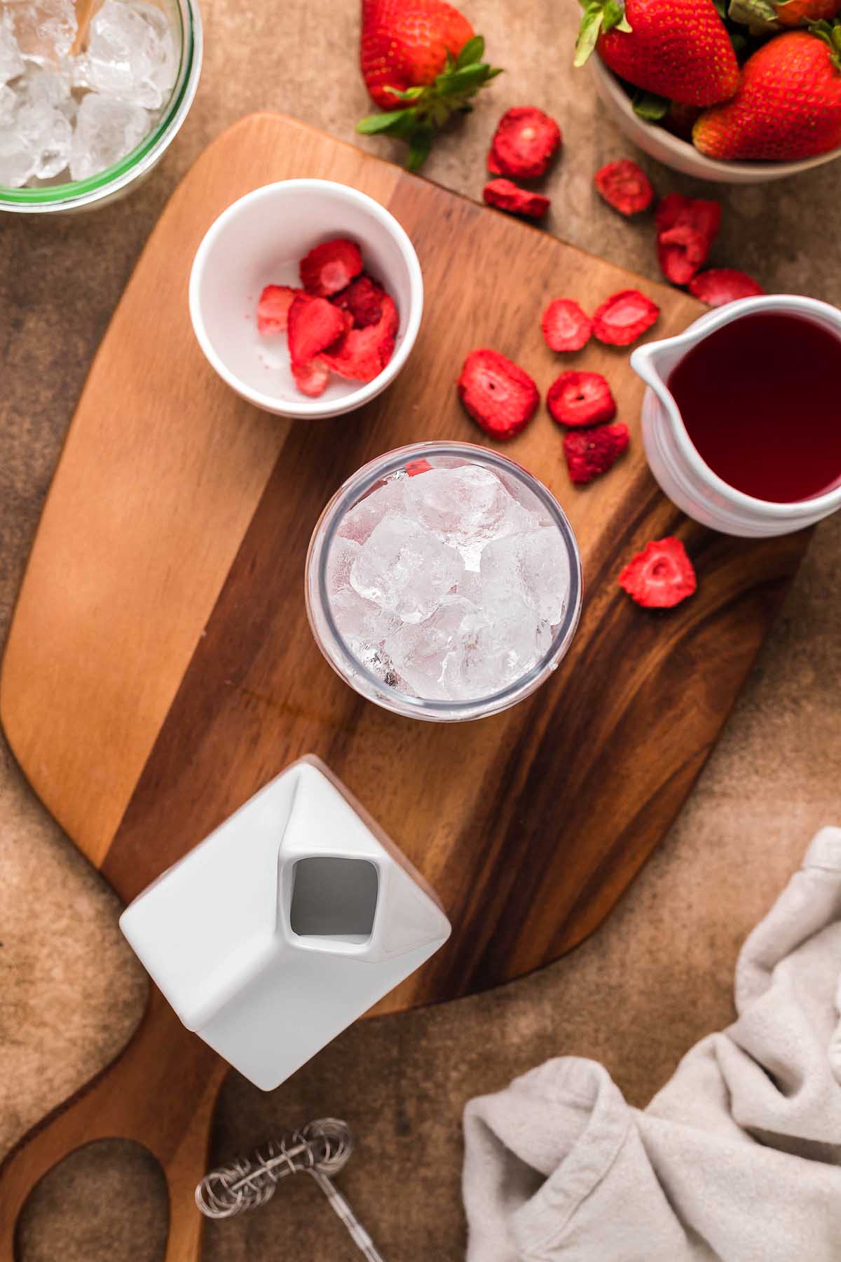 Wooden board with a glass of ice, a white pitcher, sliced strawberries, and a cup of freeze-dried strawberry slices—perfect for crafting your own homemade Starbucks pink drink. A cloth and a whisk are nearby.