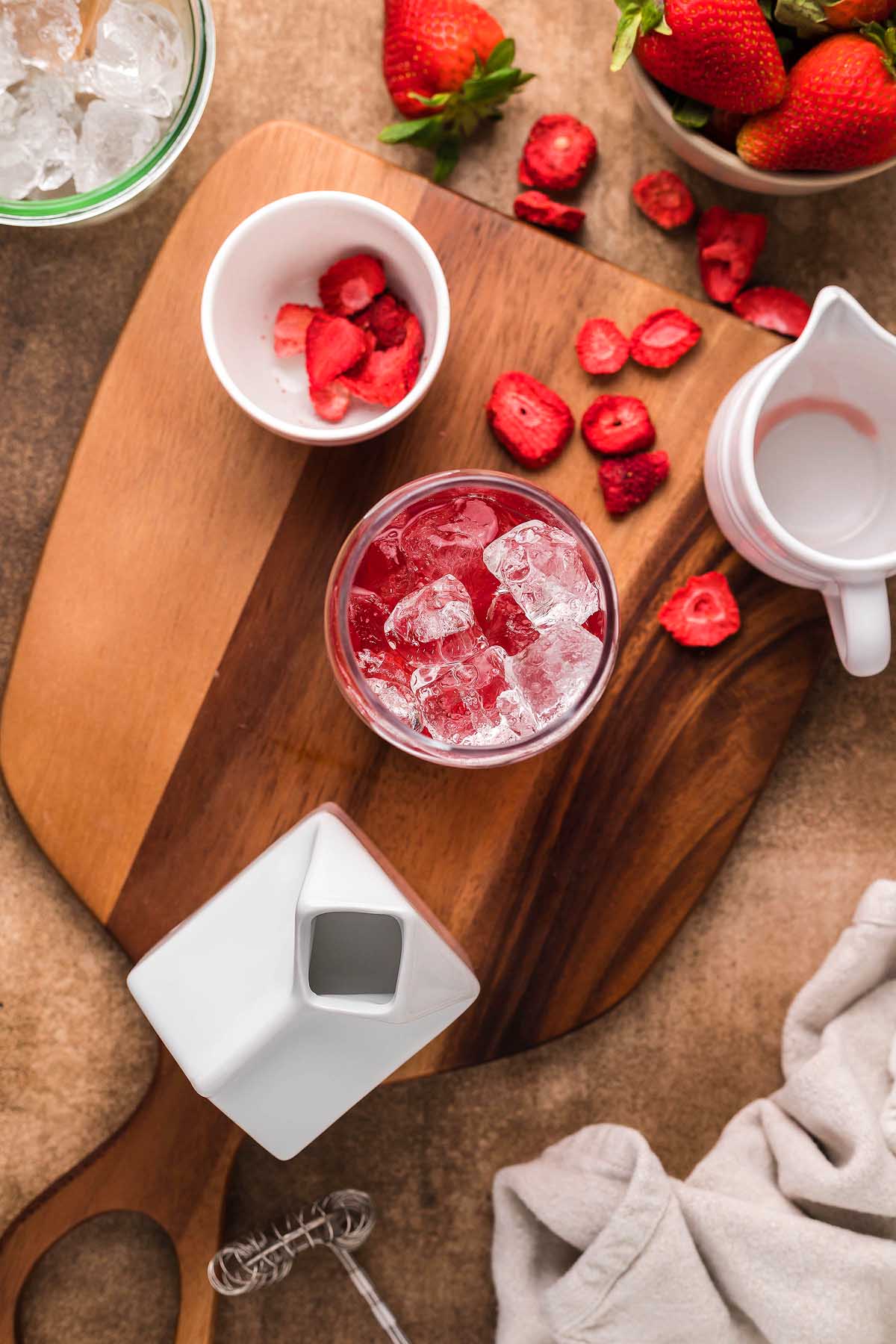 A wooden board holds a glass of Starbucks Copycat Pink Drink with ice, surrounded by sliced freeze-dried strawberries, an empty pitcher, a bowl, and a cloth.