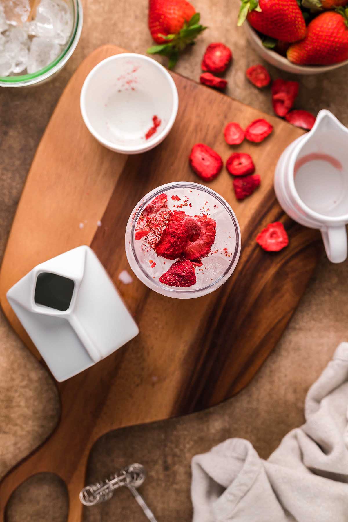 A glass filled with ice and sliced strawberries sits on a wooden board, alongside a pitcher, a small white jar, and a bowl of strawberries—perfect for crafting your own Starbucks Copycat Pink Drink at home.