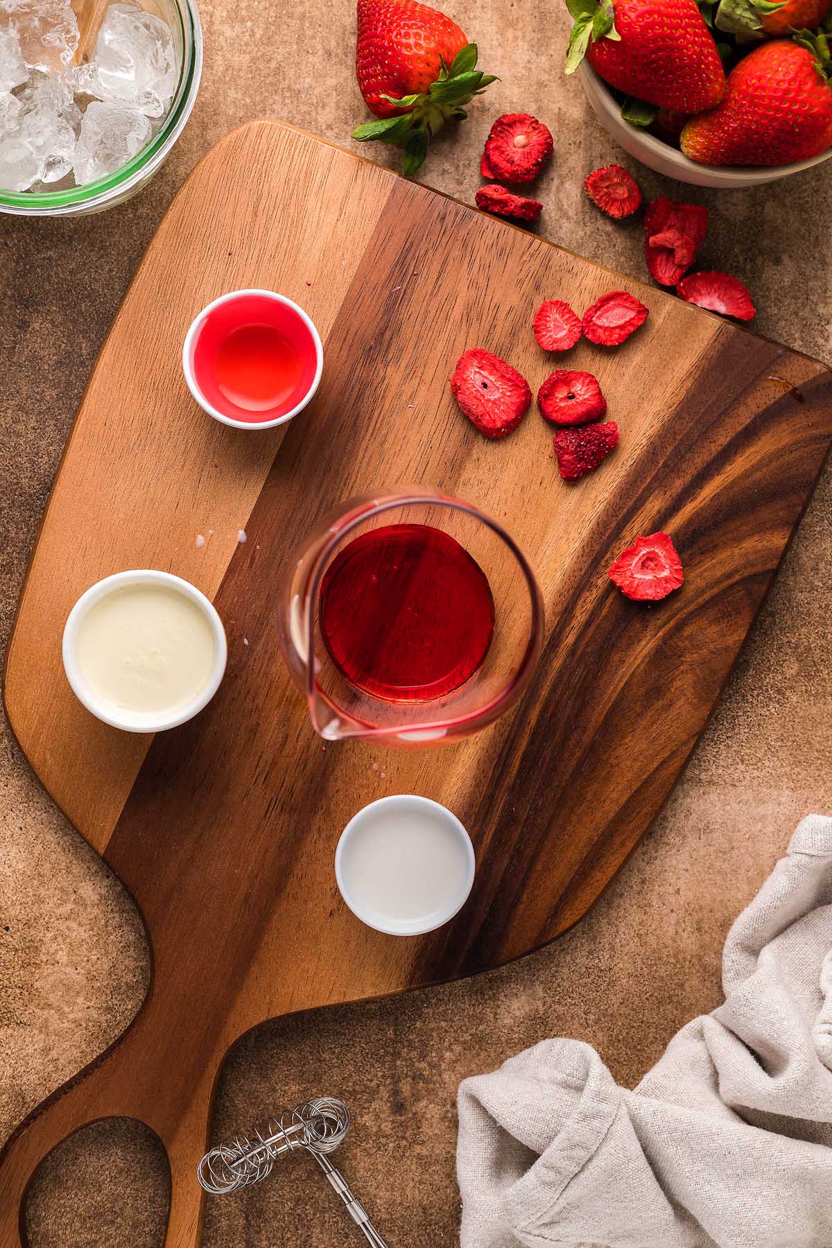 On a wooden cutting board sits a glass of tantalizing red liquid, surrounded by small bowls of white and red liquids. Sliced freeze-dried strawberries, a bowl of whole berries, and crushed ice add flavor to this inviting scene—perfect for crafting your own Starbucks Pink Drink copycat recipe at home.
