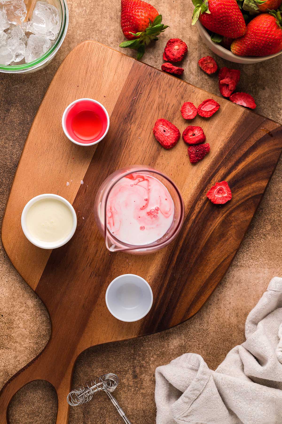 Cutting board with freeze-dried strawberries, a small pitcher of a Starbucks Copycat Pink Drink, three small cups containing white, clear, and red liquids, an ice bowl, and a white cloth on the side.