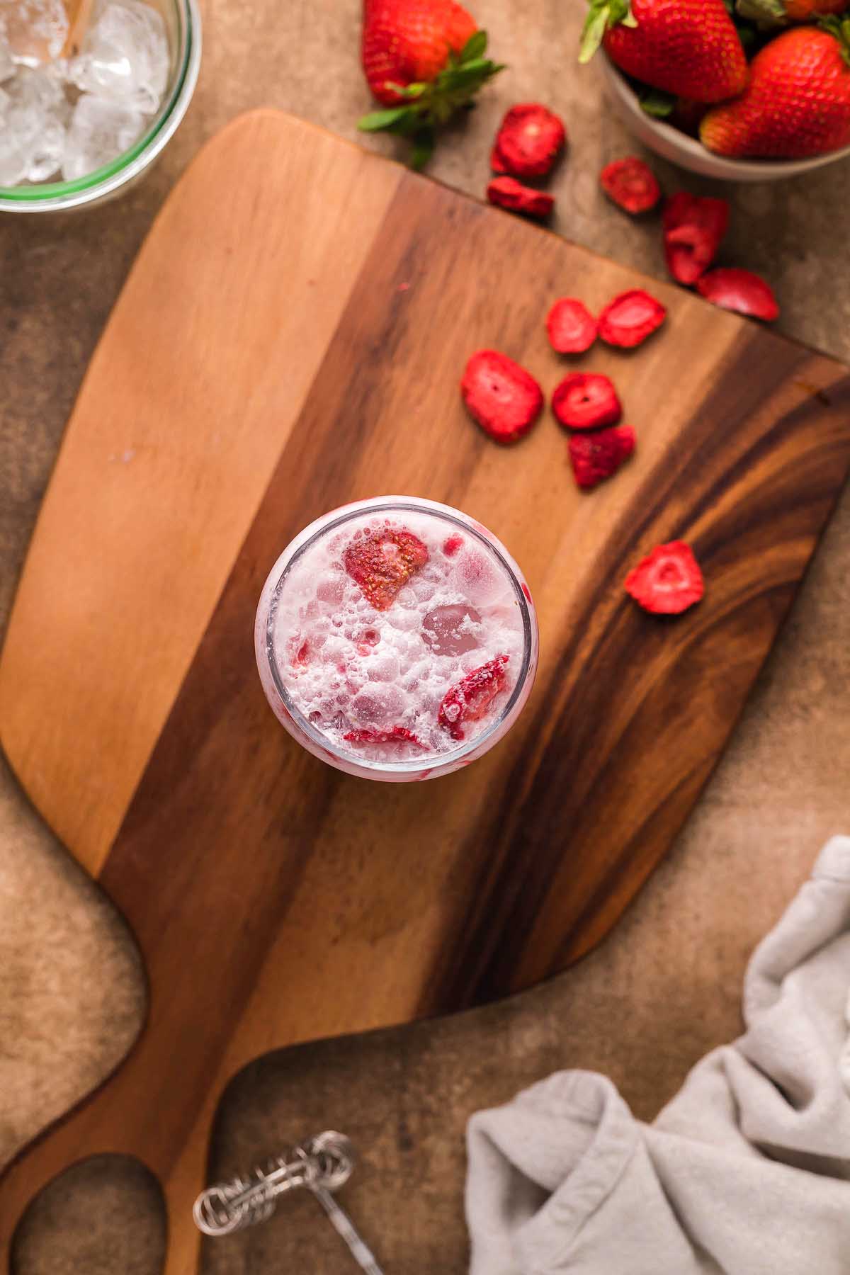 A glass of pink iced drink with sliced freeze-dried strawberries sits on a wooden board, surrounded by fresh strawberries, ice, and a cloth—just like the Starbucks Pink Drink copycat recipe.