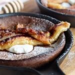 A gluten free dutch baby skillet pancake topped with caramelized banana, powdered sugar, and whipped cream on a wooden board.