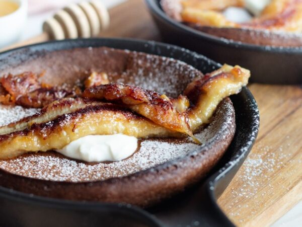 A gluten free dutch baby skillet pancake topped with caramelized banana, powdered sugar, and whipped cream on a wooden board.