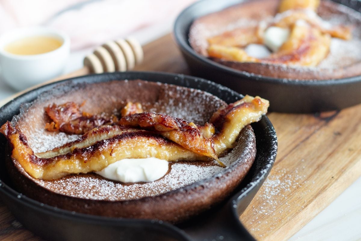 Close-up of a Chocolate Dutch Baby pancake with caramelized banana, powdered sugar, and cream in a small skillet.