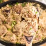 A wooden spoon holding creamy meatballs over a dish of meatballs in a creamy sauce, garnished with herbs—a delicious scene straight from a Swedish meatball recipe.