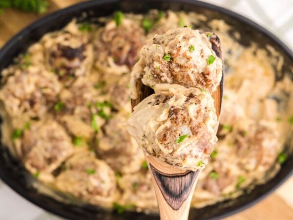 A wooden spoon holding creamy meatballs over a dish of meatballs in a creamy sauce, garnished with herbs—a delicious scene straight from a Swedish meatball recipe.