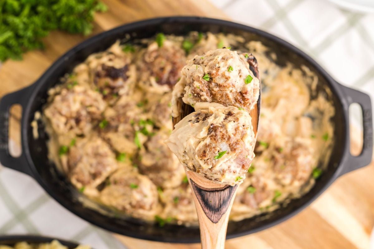 A wooden spoon holds creamy meatballs above a dish filled with more meatballs in a luscious sauce—perfect for anyone craving a copycat Ikea Swedish meatballs recipe.