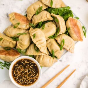 A plate of gluten free potstickers topped with sliced green onions, dipping sauce, and sesame seeds sits on a marble surface.