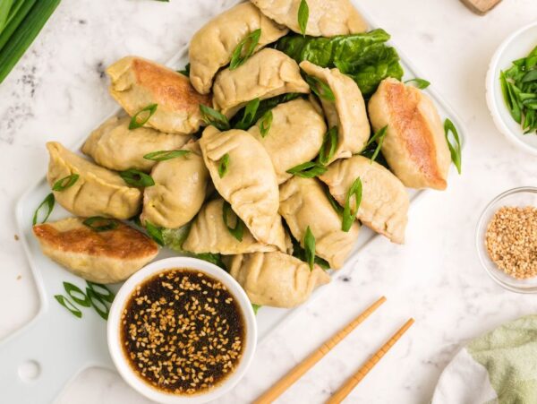 A plate of gluten free potstickers topped with sliced green onions, dipping sauce, and sesame seeds sits on a marble surface.